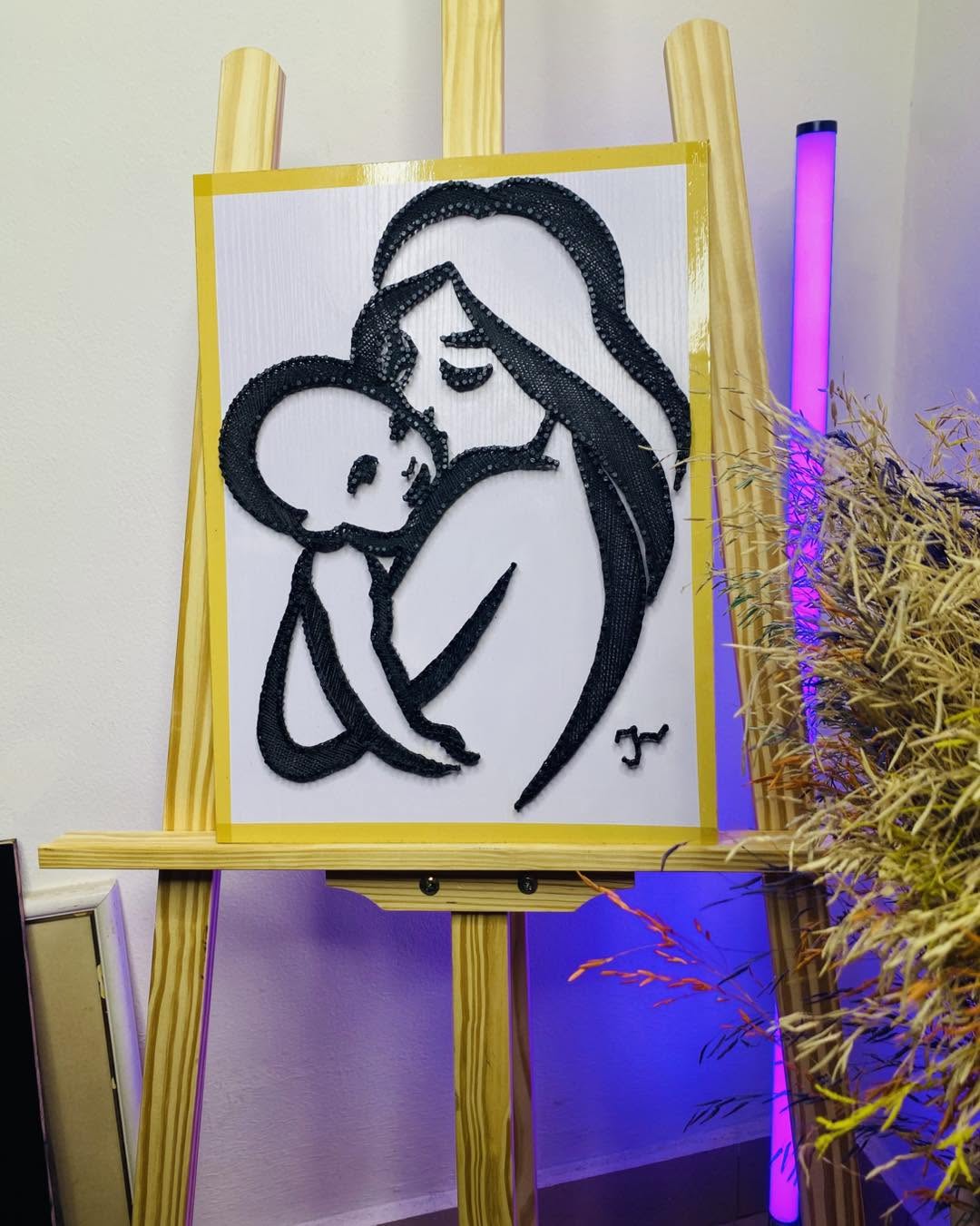 Tableau en fils noirs représentant une mère enlaçant son bébé, présenté sur un chevalet
