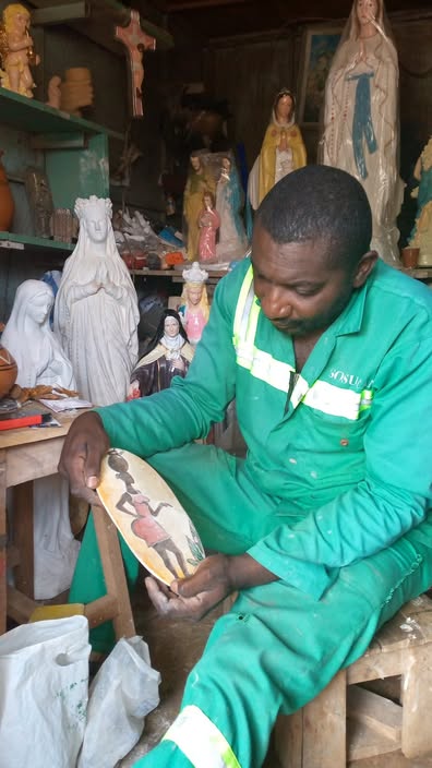 Artisan en tenue de travail verte tenant une pièce ovale peinte, entouré de statues en plâtre dans son atelier